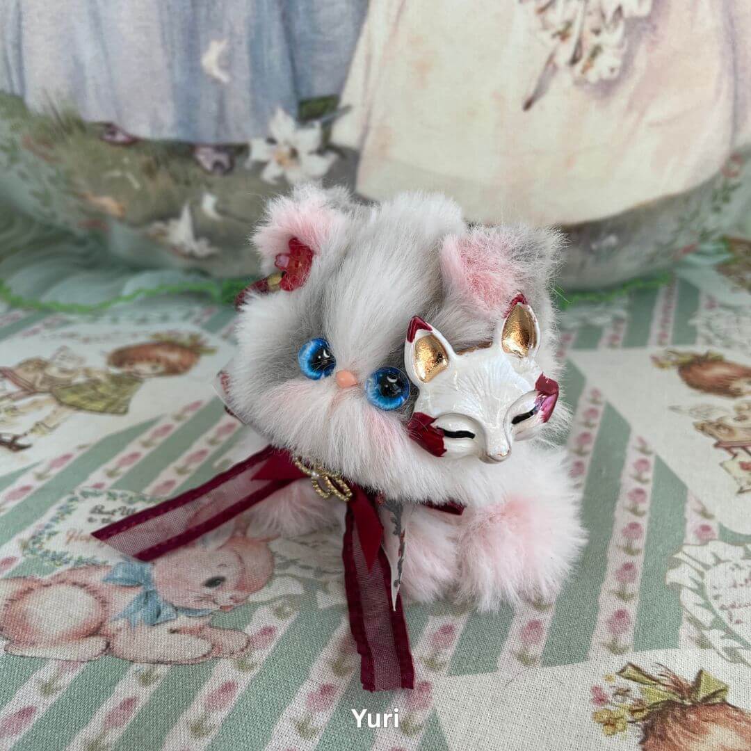 Handmade plush kitten with blue eyes wearing a fox mask and burgundy ribbon from the Cosplay Kitten Collection on a patterned background.