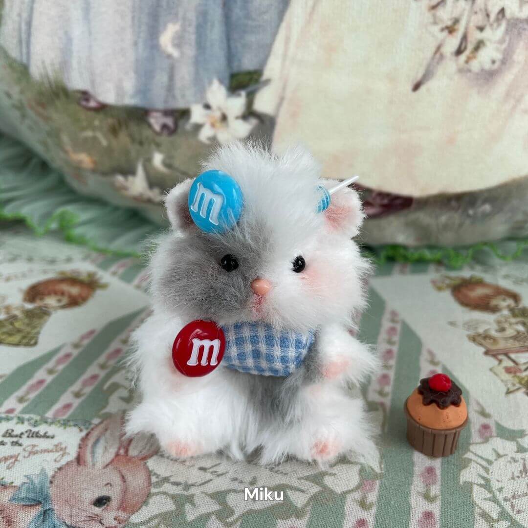 Handmade white and grey fluffy hamster figurine with blue gingham bow, decorated with colorful candy accents on a patterned fabric backdrop.