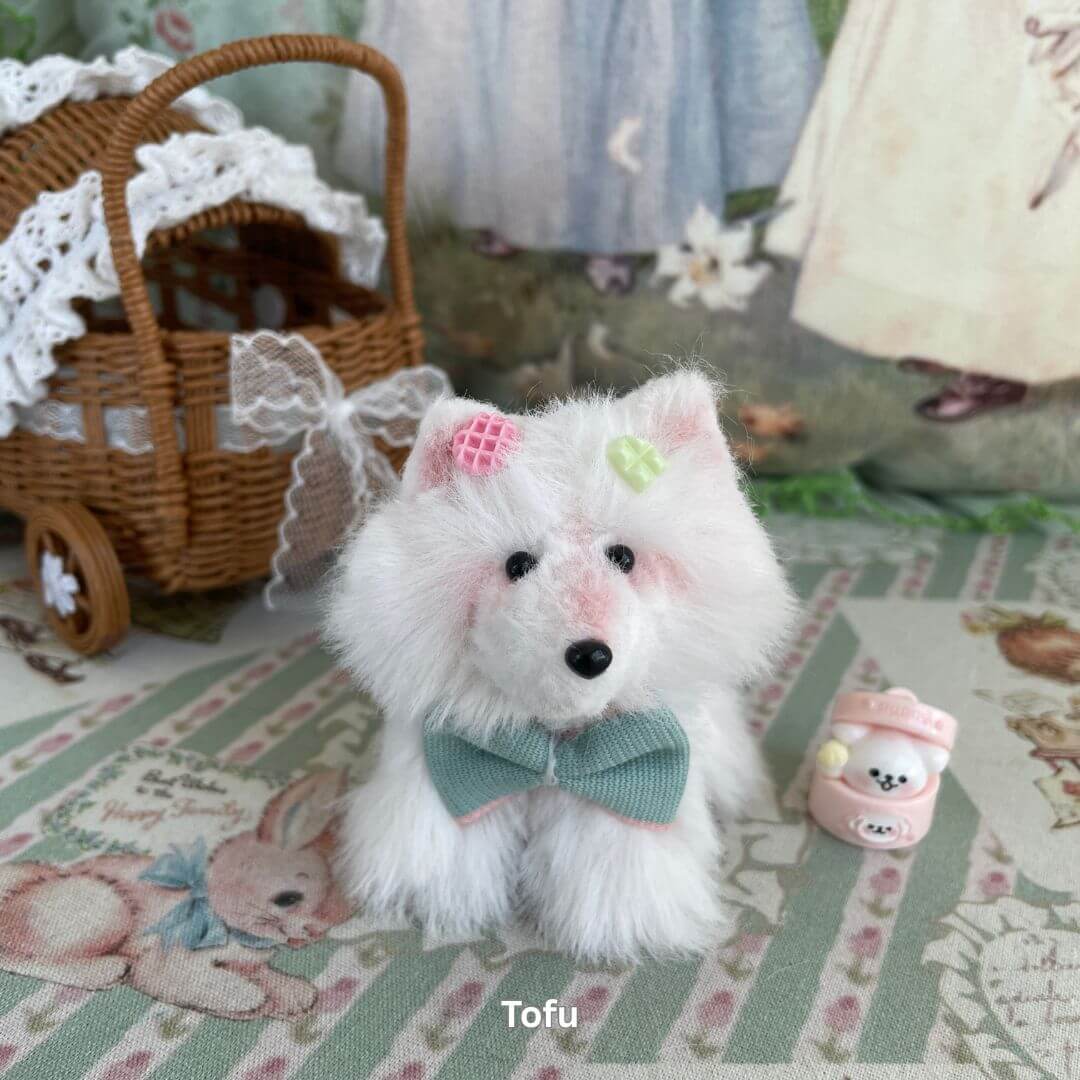 Handmade puppy plush toy 'Tofu' with white faux fur, pink bow, and green ribbon, posed beside a decorative cart and pastel accessory.