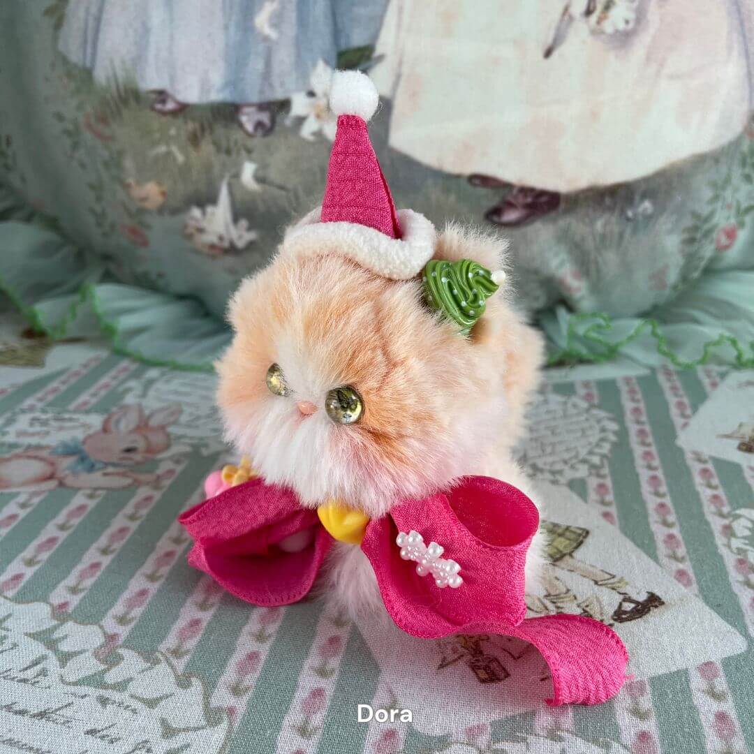 Handmade plush kitten from the Fairy Paws Collection, featuring festive hat, pink bow, and intricate detailing on a patterned background.