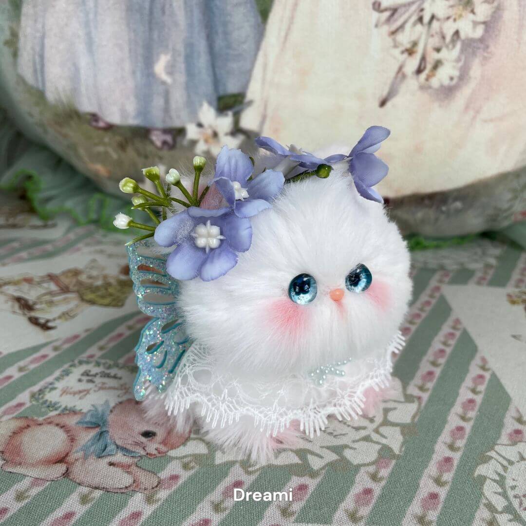 Handmade plush kitten with fairy wings, blue eyes, and floral headpiece from the Fairy Paws Collection, on a floral background.