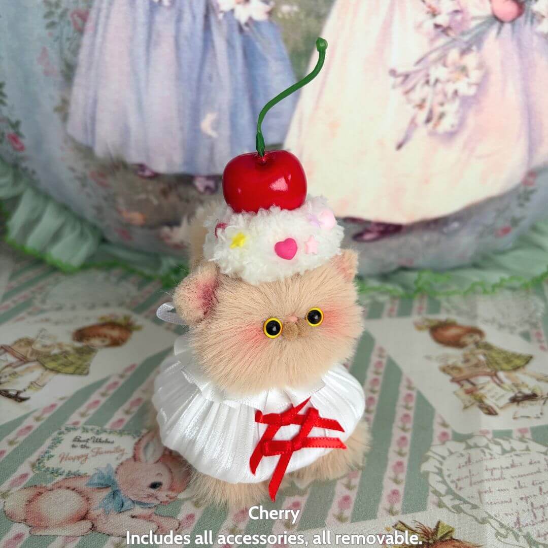 Miniature kitten with fluffy beige fur wearing a dessert-themed outfit and cherry hat, part of Sweet Treat Kitties collectible.