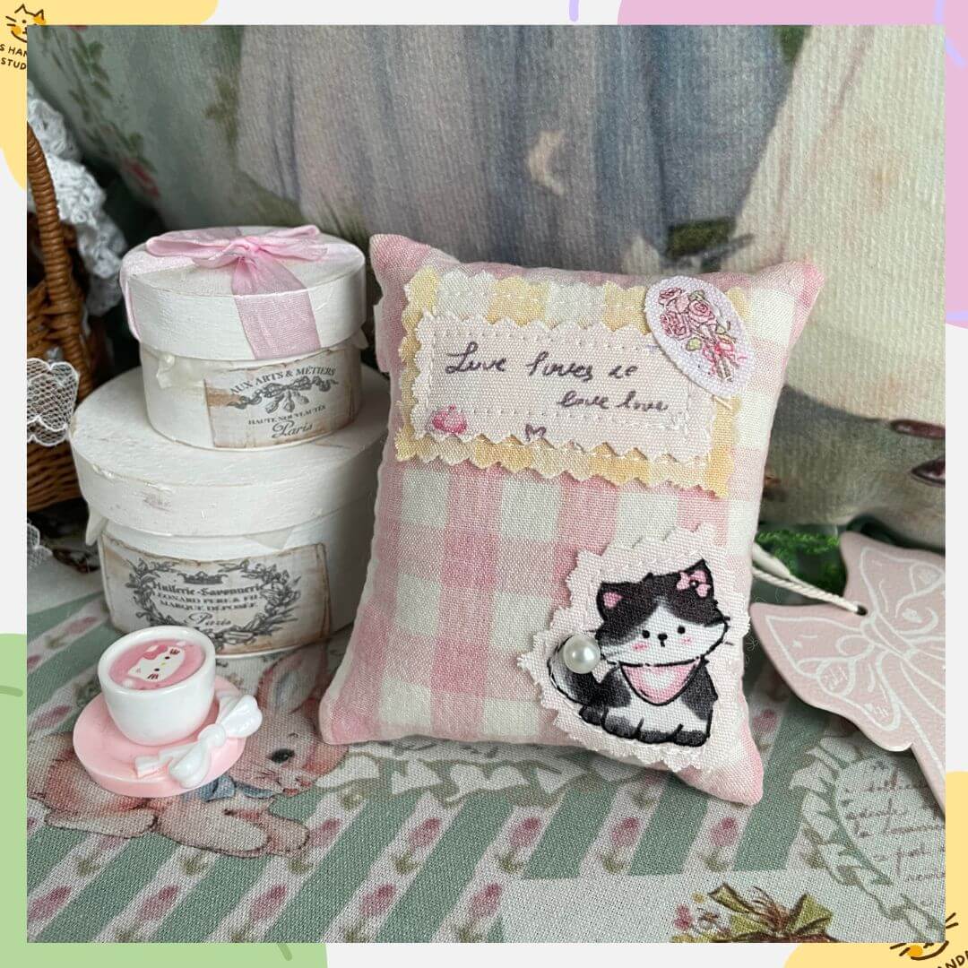 Mini handmade pillow bag charm with kitten design, crafted for dollhouse decor, featuring 'live loves' embroidery and checkered fabric.