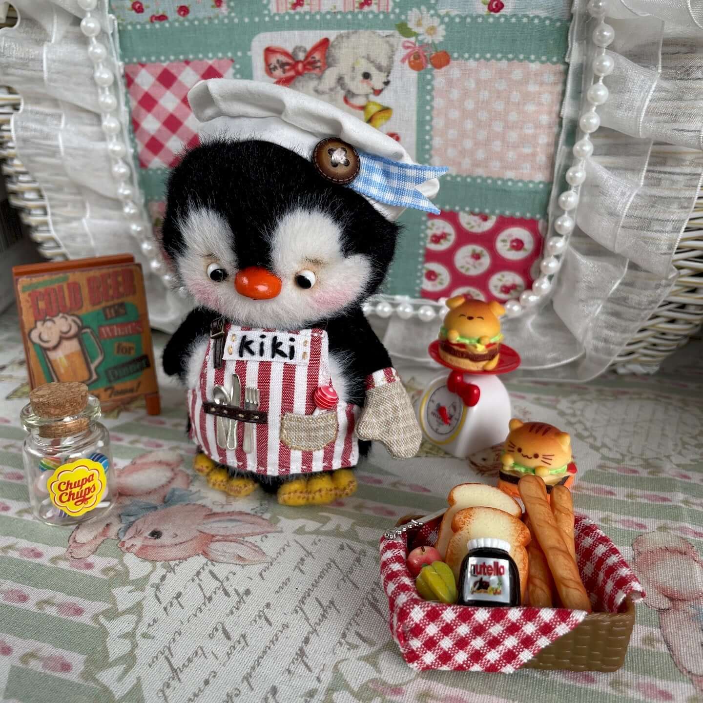 Mini Chef Penguin with Movable Joints in striped apron surrounded by miniature kitchen accessories and a basket of toy food items.