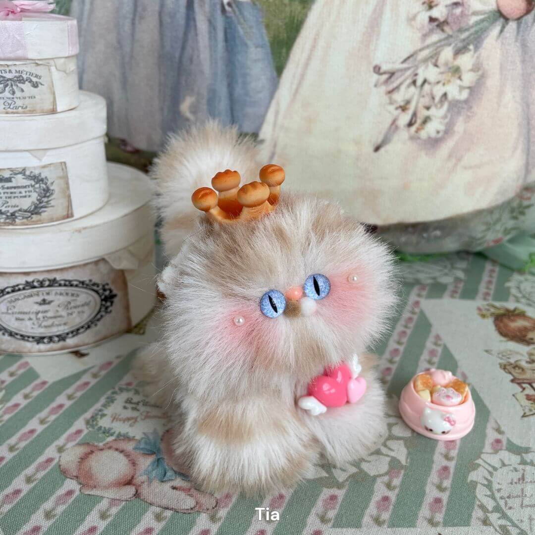 Handmade fluffy kitten plush toy with blue eyes, holding a heart. Adorned with crown and surrounded by decorative items. Soft pastel colors.