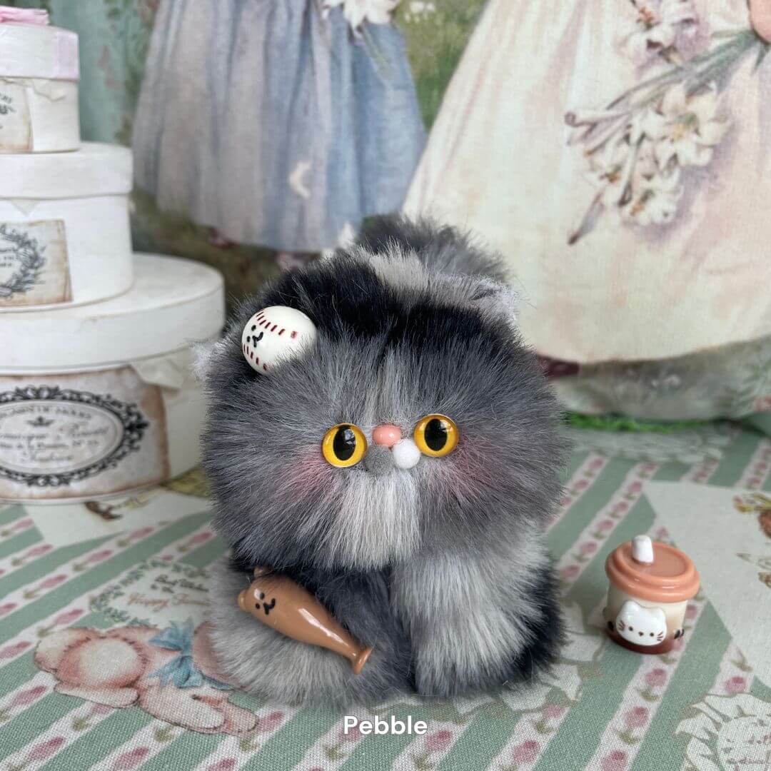 Fluffy handmade plush kitten toy in grey and black with yellow eyes, adorned with a small cone decoration, sitting on a floral backdrop.