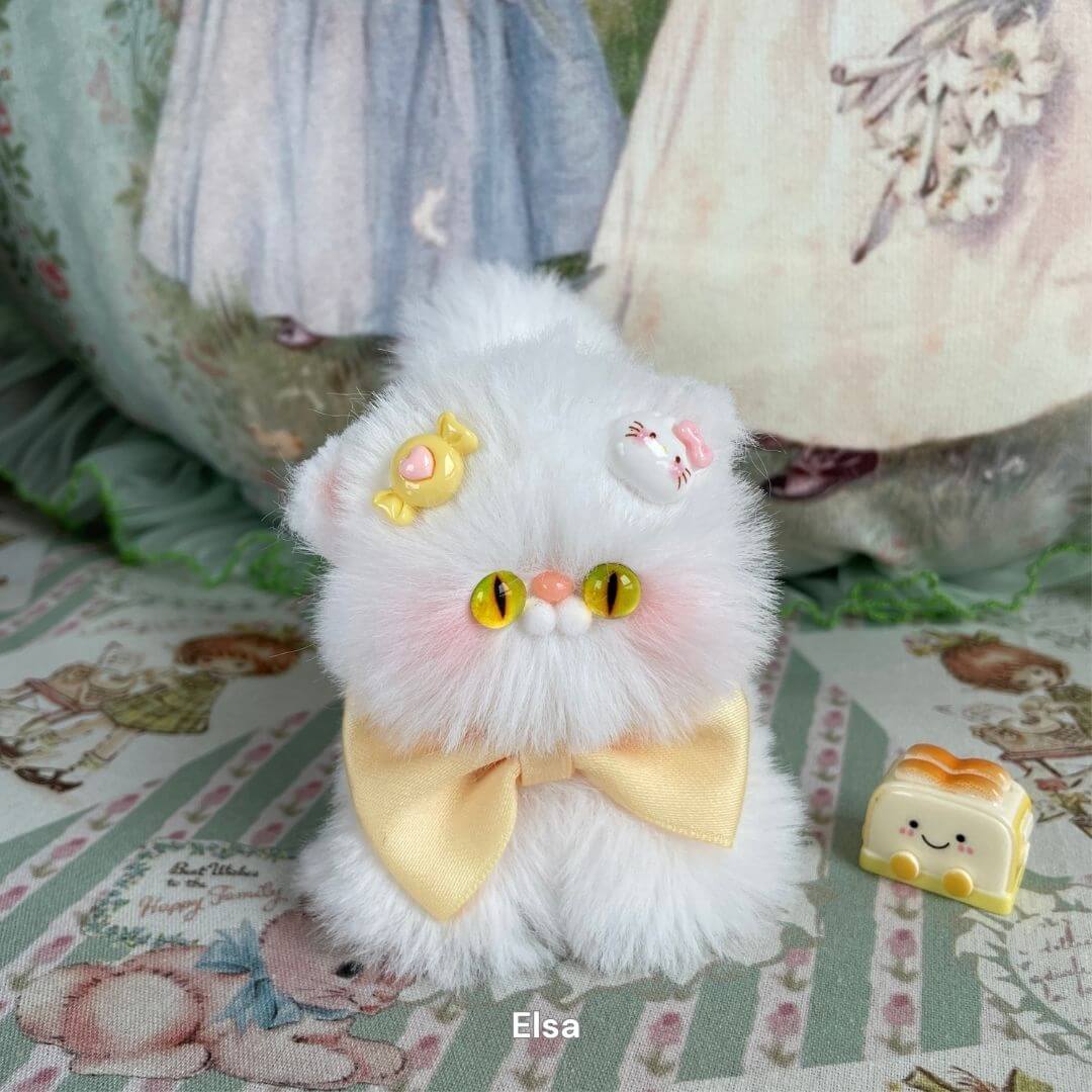 Fluffy white plush kitten toy with bright yellow eyes, decorated with a pastel pink candy and yellow bow on a floral fabric background.