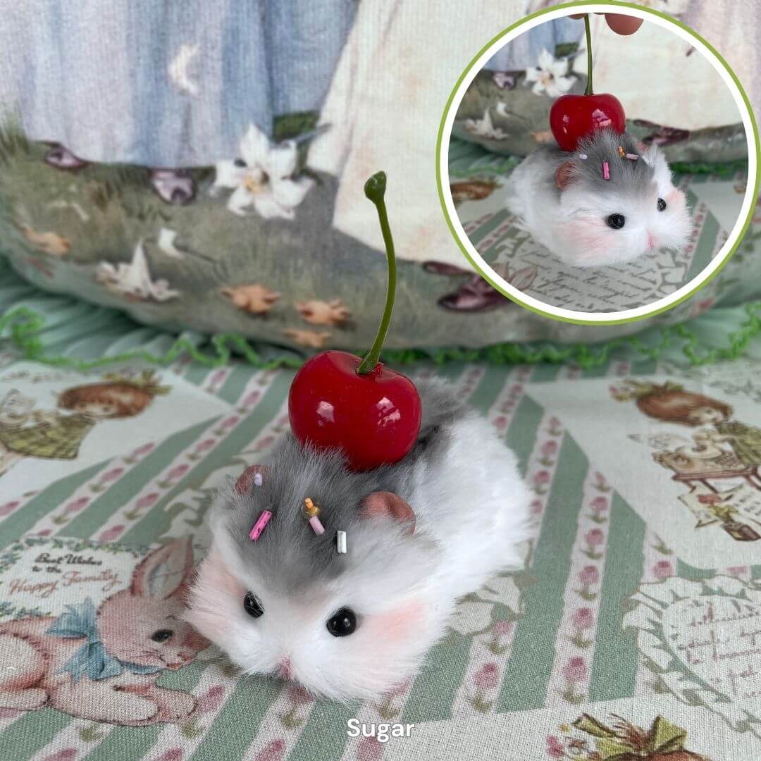 Handmade lying hamster figurine with a cherry on its back, crafted from soft faux fur, on patterned fabric with floral decorations.