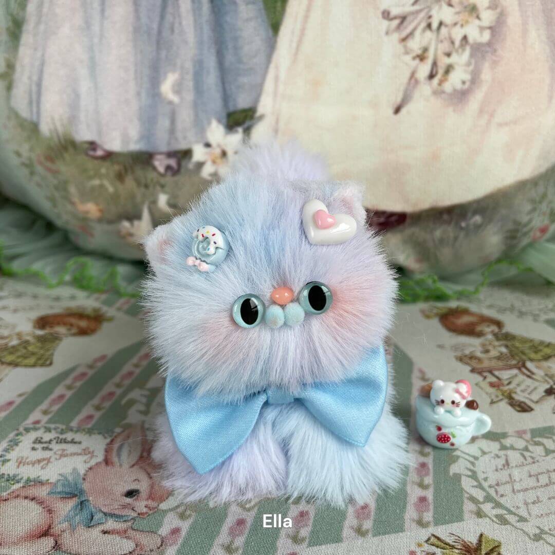Handmade plush pastel kitten with bow from Fluffy Kitten Collection, featuring soft faux fur and decorative badges in a whimsical setting.