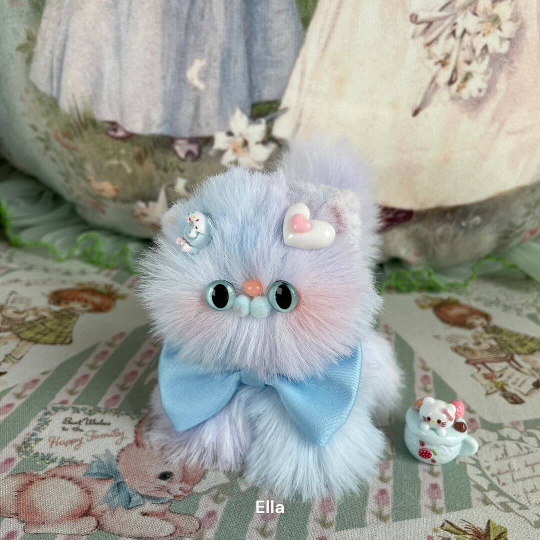 Fluffy pastel kitten plush toy with blue bow and decorative heart and charm, named Ella, on a vintage fabric background.