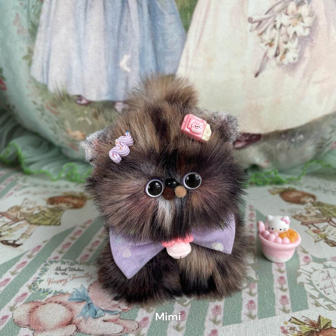Handmade fluffy kitten plush toy with purple bow and candy decoration, part of the Fluffy Kitten Collection, on floral background.