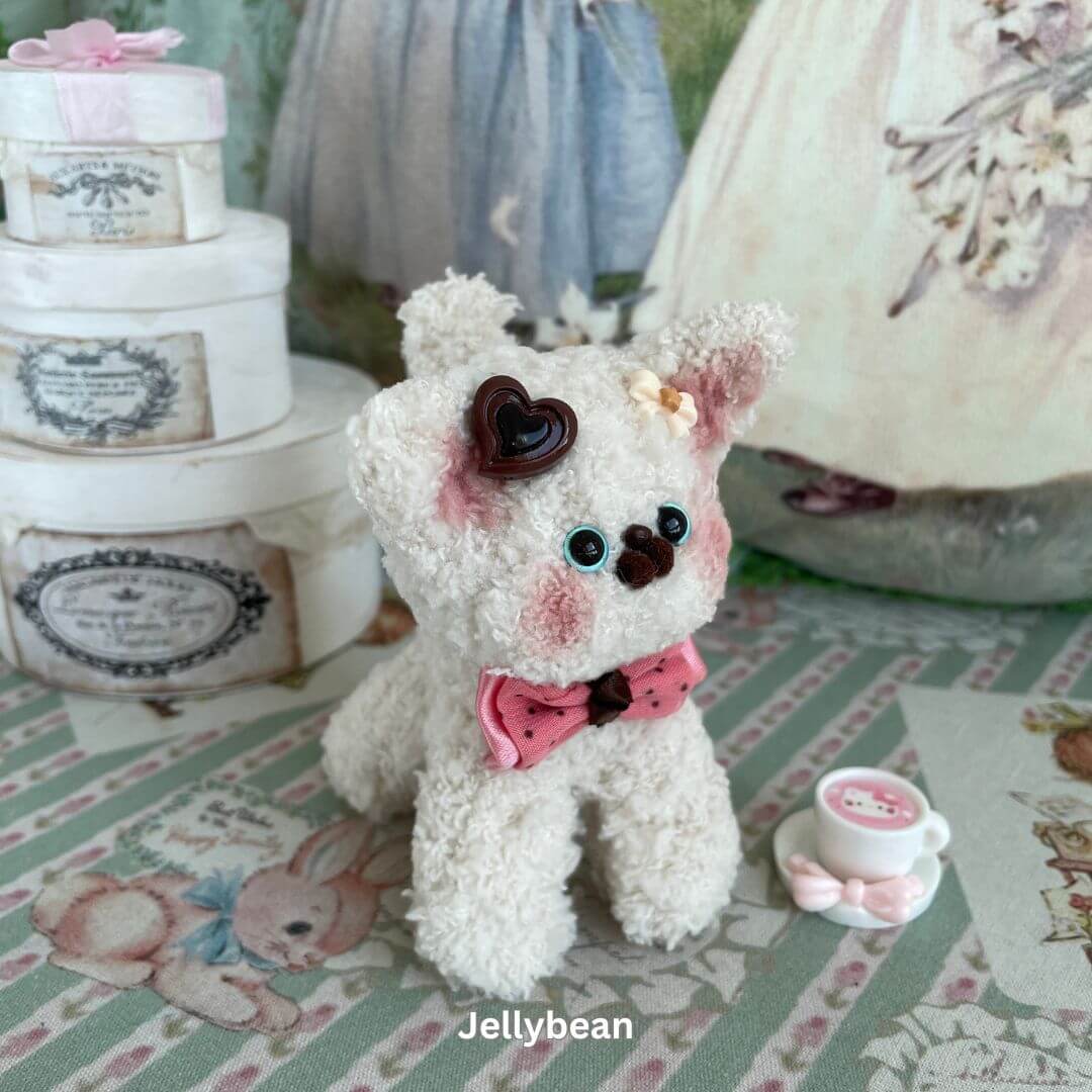 Handmade plush toy puppy with heart decoration and pink bow, set against a whimsical background with decorative boxes and a teacup.