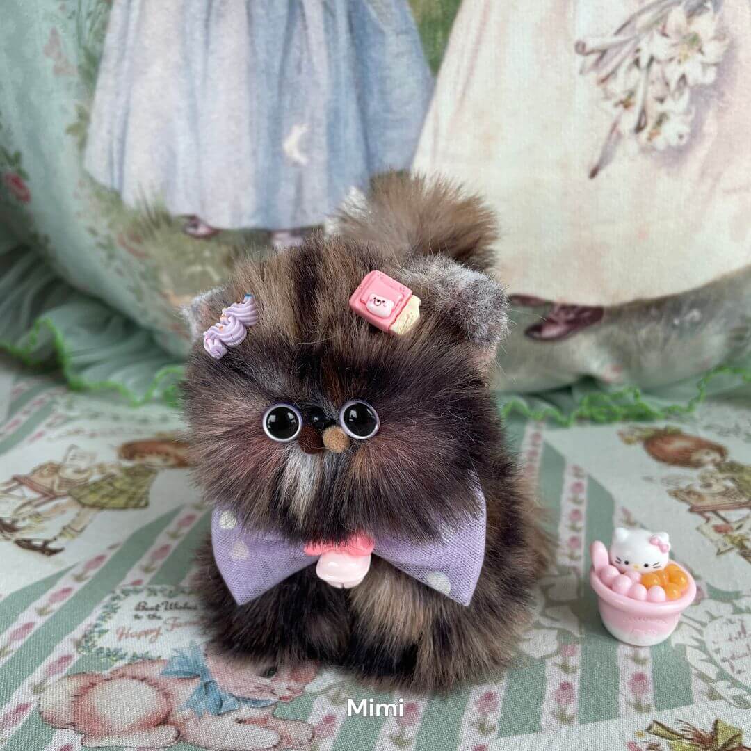 Handmade fluffy kitten plush toy with purple bow and accessories, part of Fluffy Kitten Collection, on a decorative background.