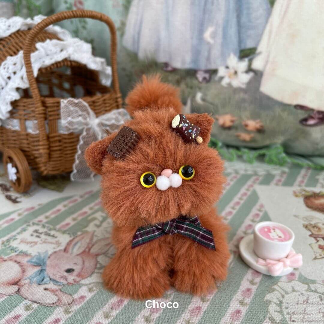 Handmade brown kitten plush toy with dessert-themed accessories, including a cookie bow and whipped cream clip, on a patterned surface.