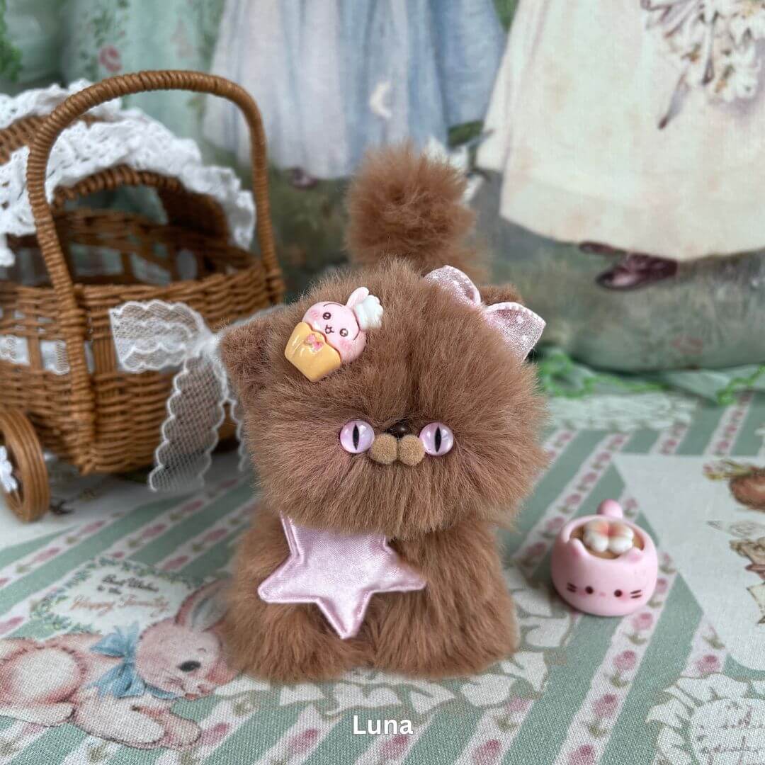 Handmade brown kitten plush toy with cupcake accessory and pink star bow, styled in a whimsical setting with wicker basket.