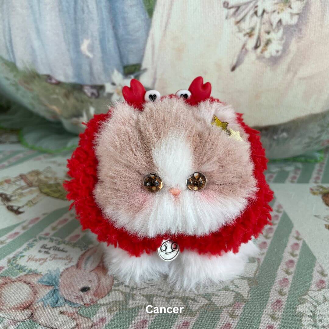 Handmade Cancer zodiac kitten plush toy with red faux fur, soft horns, and charm accessory on a decorative fabric background.
