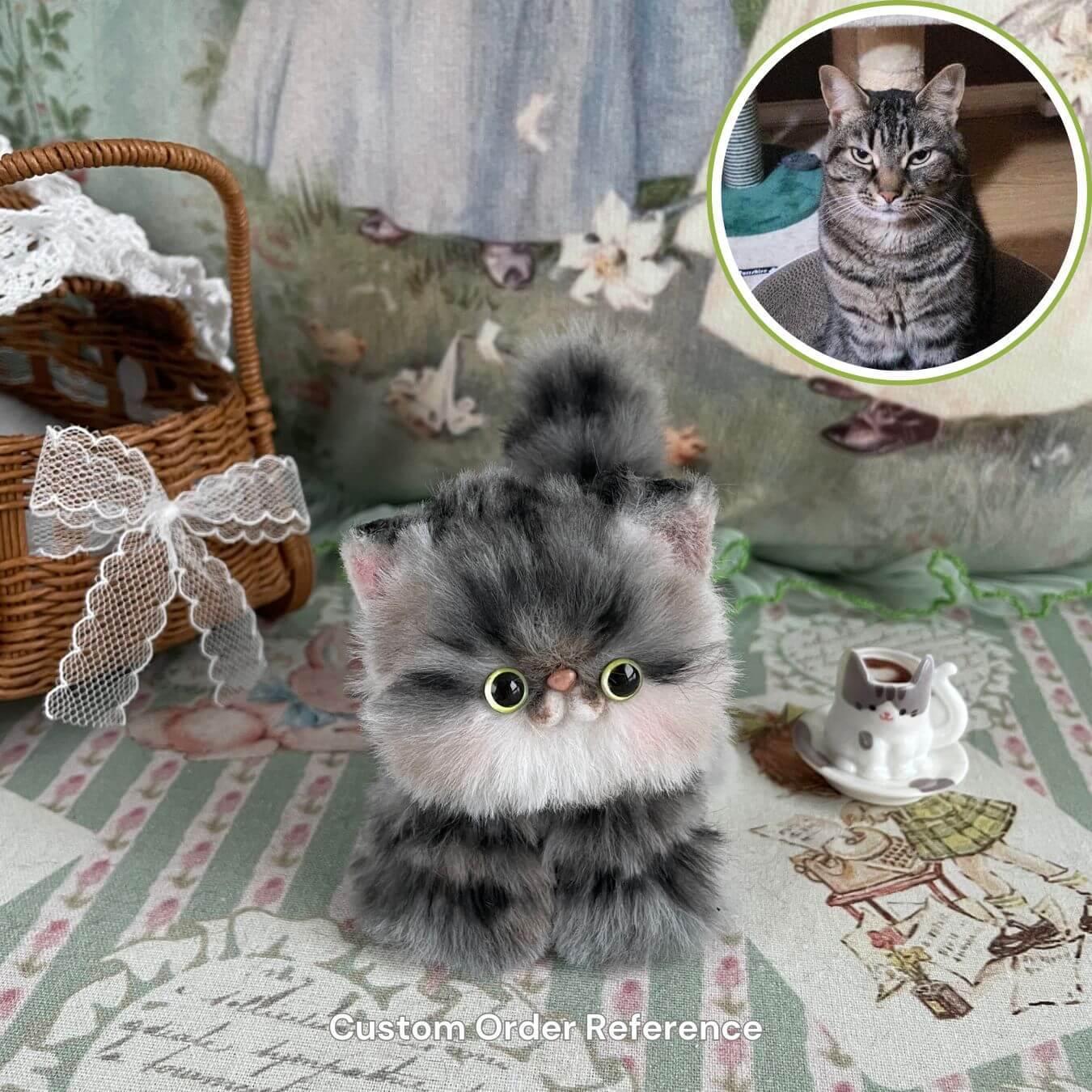 Handmade fluffy kitten plush toy with a reference photo of a real cat, surrounded by lace basket and teacup decoration.