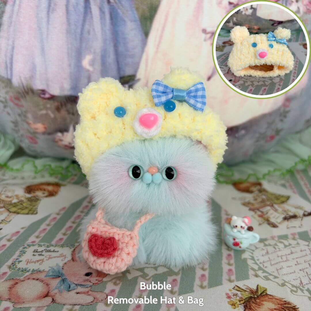 Fluffy Hoodie Kittens: cute blue kitten figurine wearing a yellow bear hat and pink bag, surrounded by pastel decor.