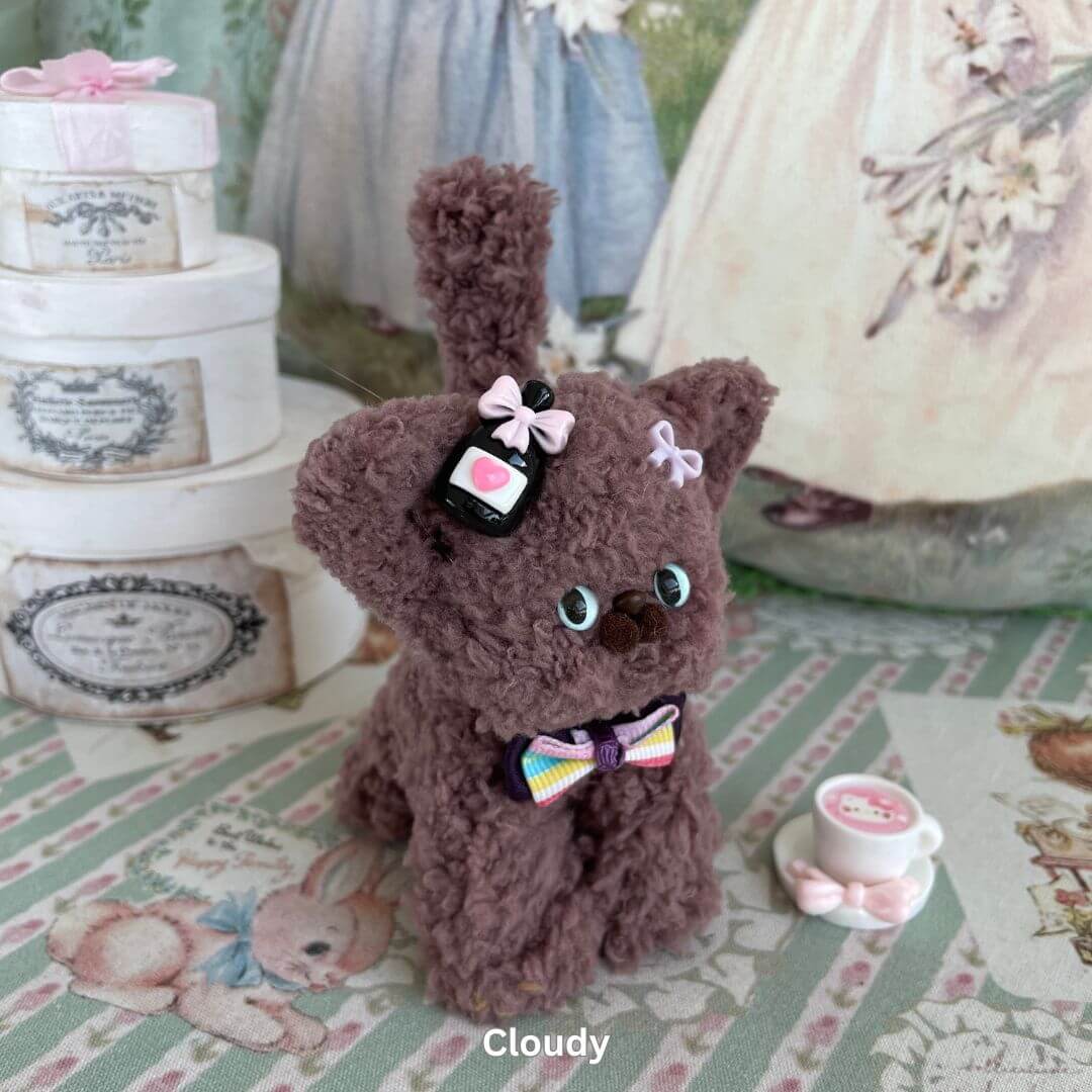 Handmade Devon Rex plush kitten toy with bows, displayed with decorative boxes and a teacup, on a striped tablecloth.