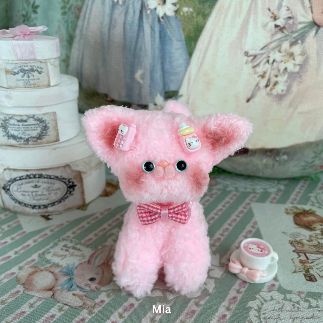 Pink plush Devon Rex kitten toy with big ears, handcrafted from faux fur, wearing a pink bow, displayed in a charming decorated setting.