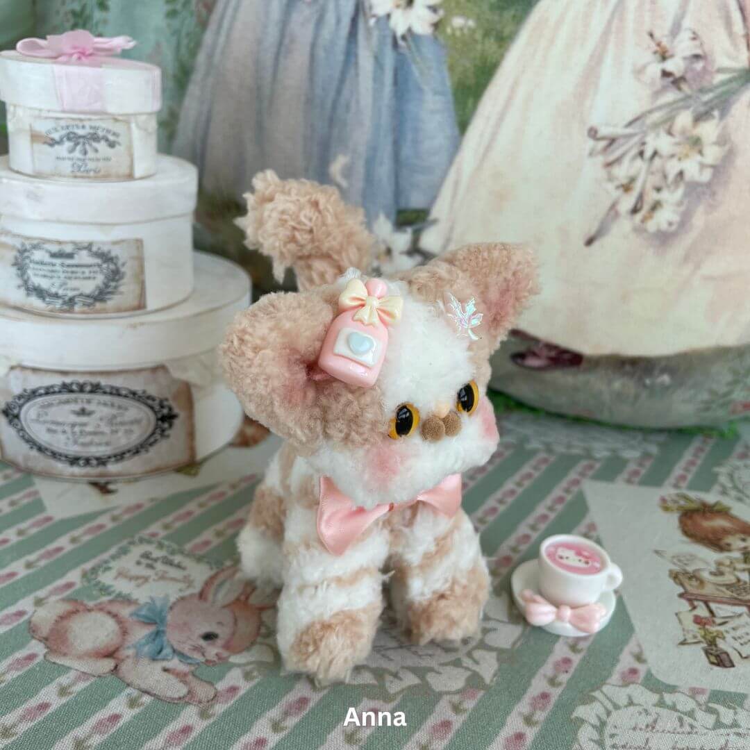 Handmade Devon Rex kitten plush with pink bow and cup accessory, inspired by the quirky charm of the real breed, sitting on patterned fabric.