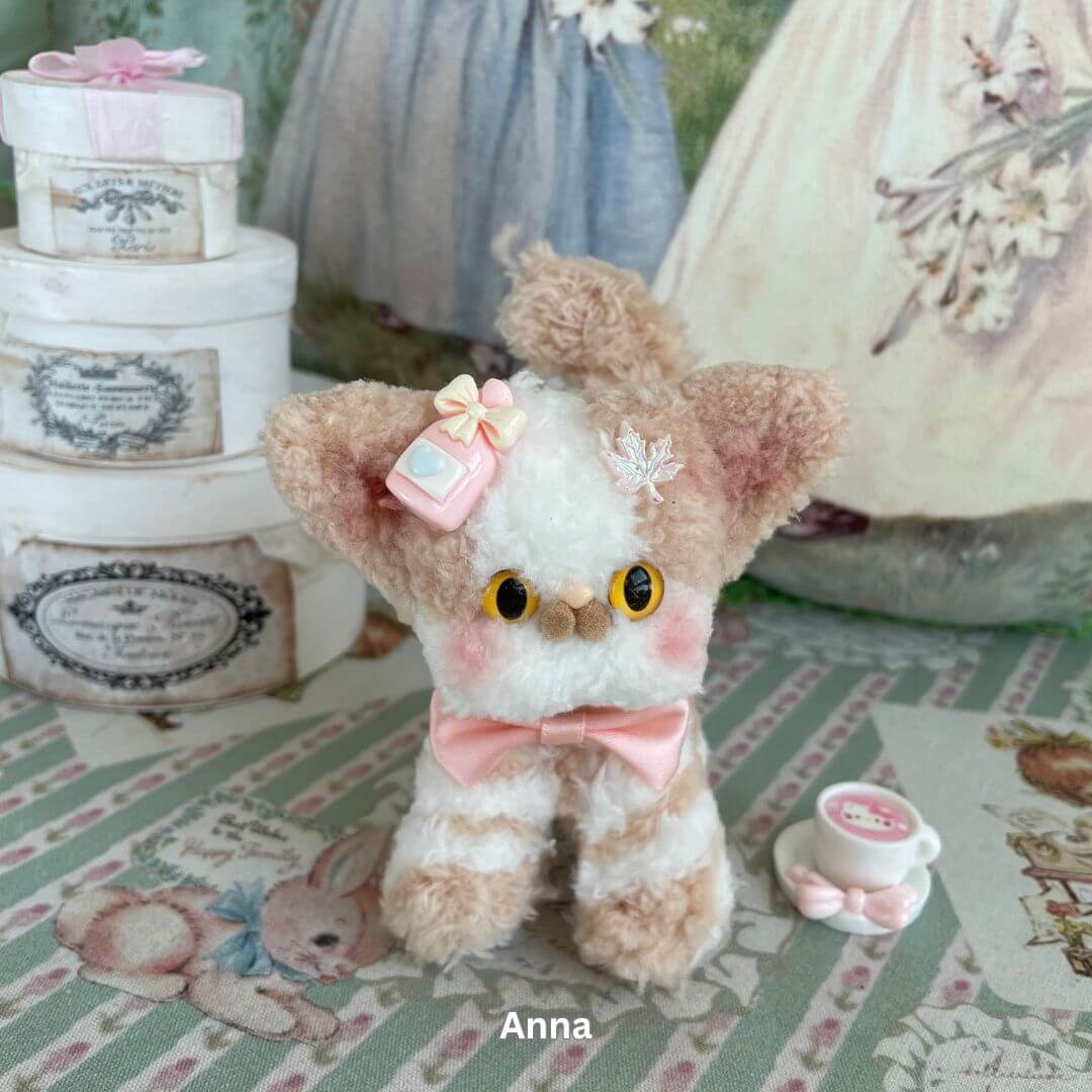 Handmade Devon Rex kitten plush toy with big eyes, a pink bow, and decorative elements, set against a vintage-style background.