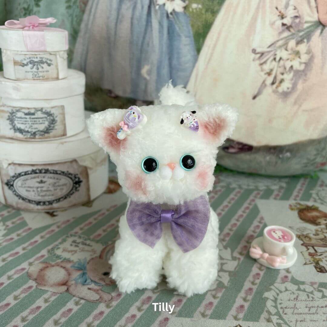 Devon Rex plush kitten with purple velvet bow and floral accessories, surrounded by vintage-style decor and tea set.