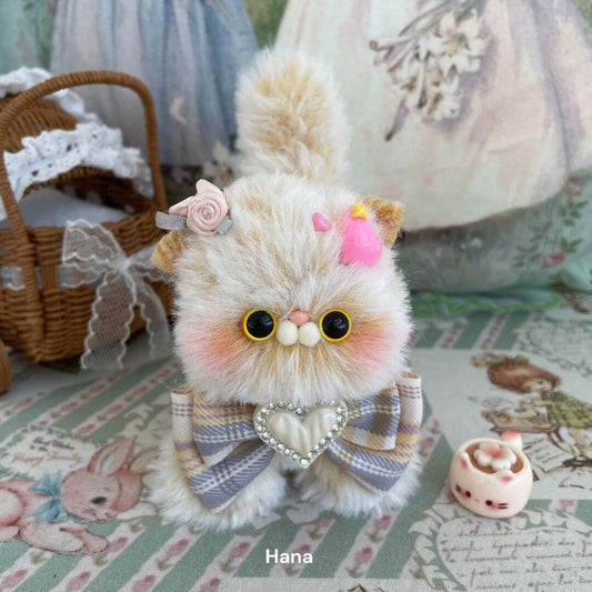 Handcrafted fluffy kitten plush toy with accessories on a decorative background, featuring a heart-shaped bow and cute embellishments.