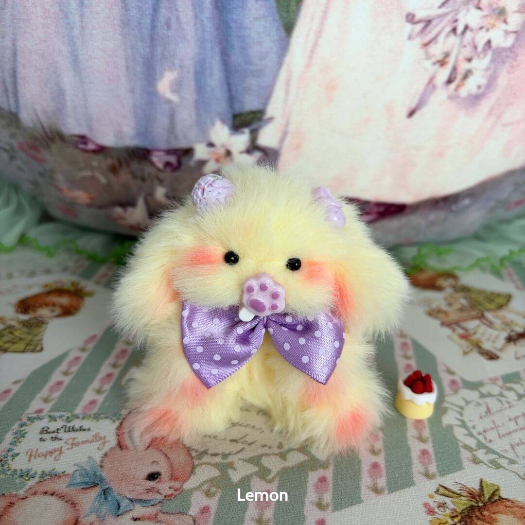 Handmade fluffy yellow bunny from the Adorable Bunny Collection, wearing a lavender bow, with pink cheeks and a tiny cake.