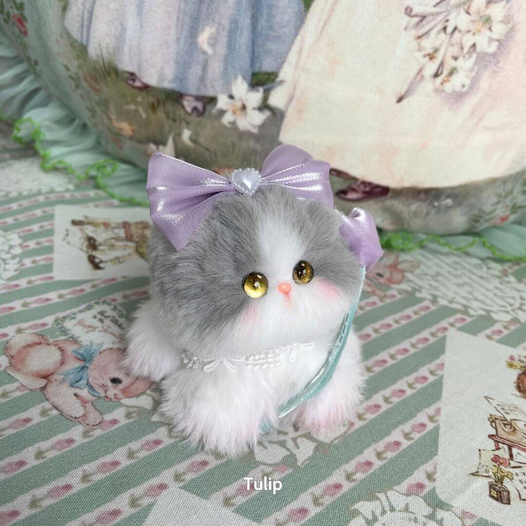A fluffy gray and white handmade kitten with a lavender bow and gentle eyes, part of the Blossom Kitten Collection.