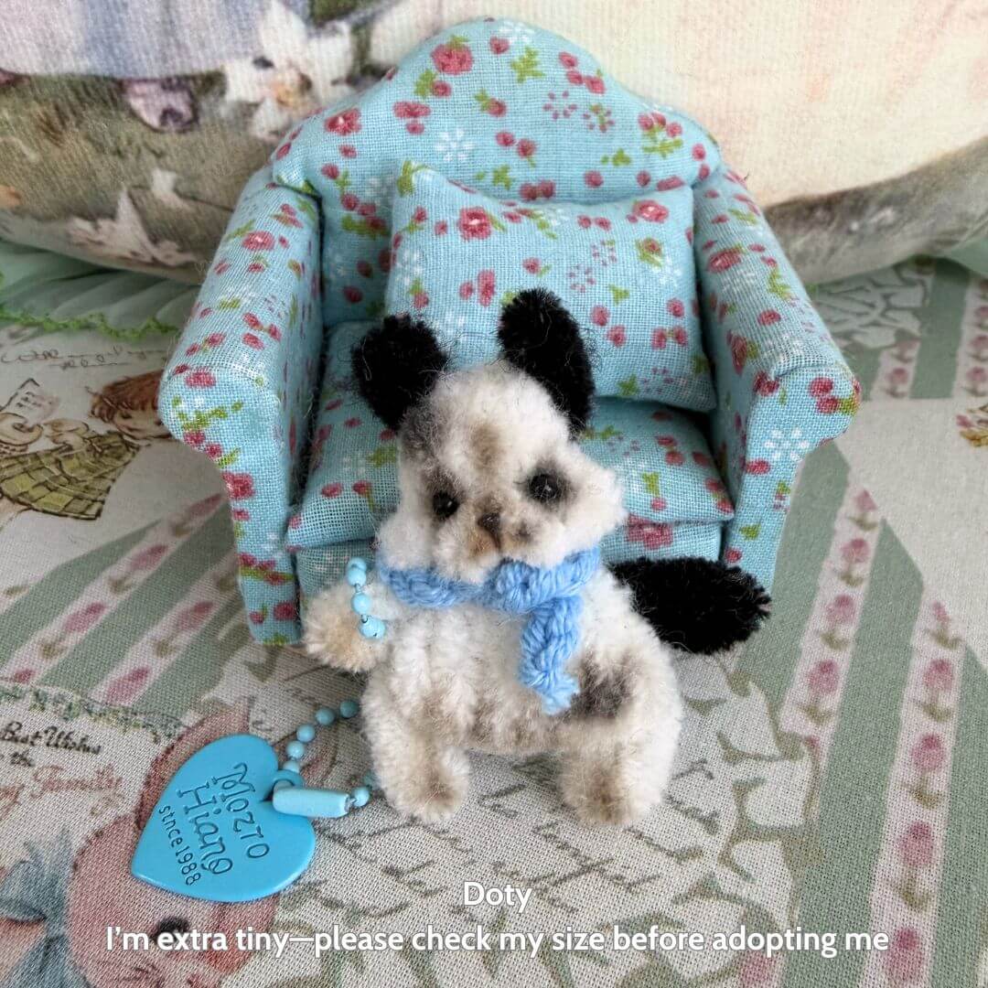 Alt Tag: Miniature Pipe Cleaner Animals featuring a fluffy kitten figurine with black and white fur, dressed in a blue scarf, sitting on a floral-patterned chair.
