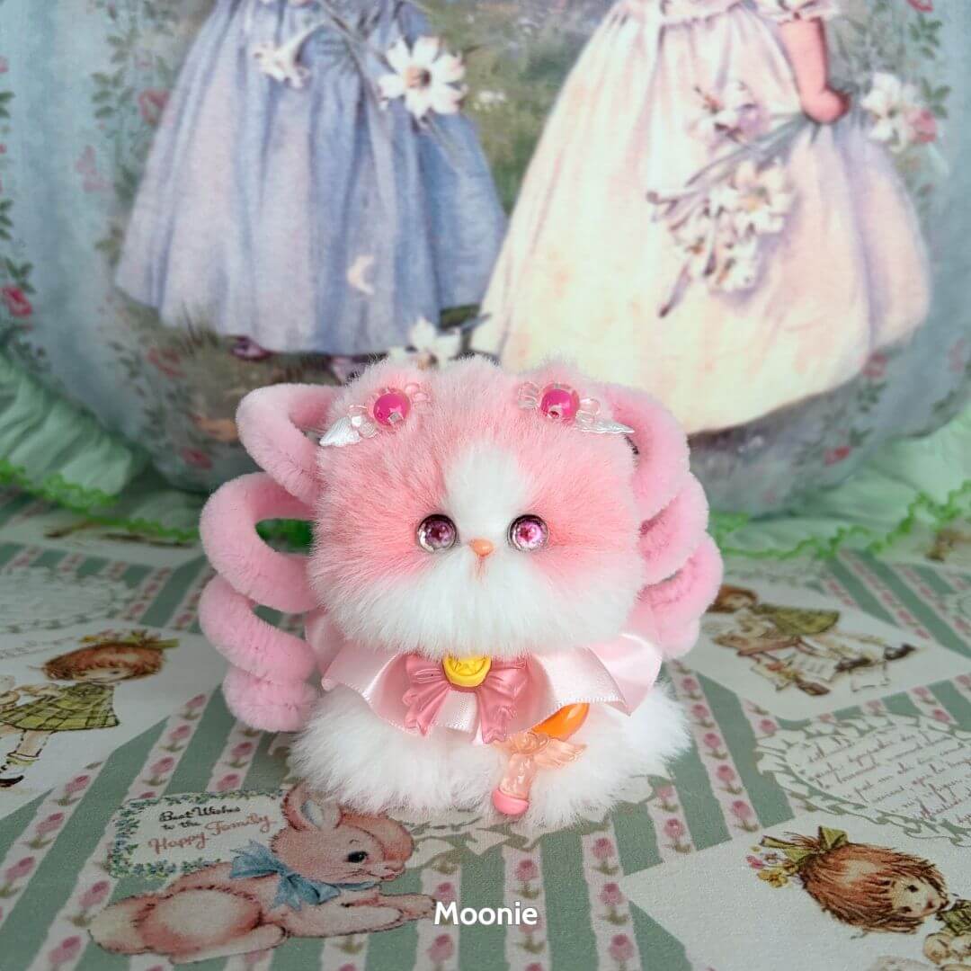 Fluffy pink handmade kitten, Moonbound Companions, with bright eyes and delicate accessories, radiating kawaii charm.