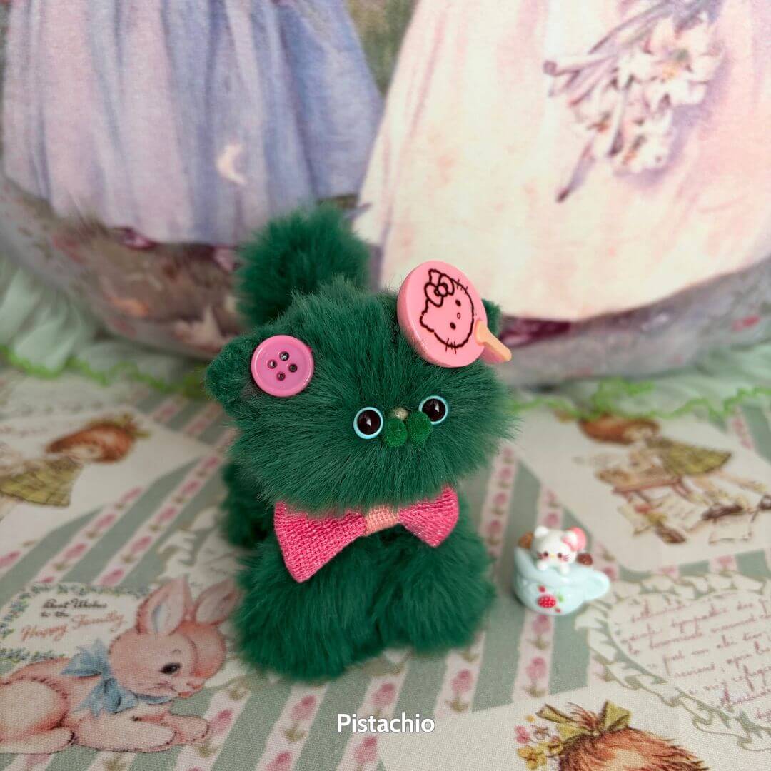Cute green handmade kitten from the Fruity Kitten Collection, adorned with pink buttons and a small bow tie, in a whimsical pose.