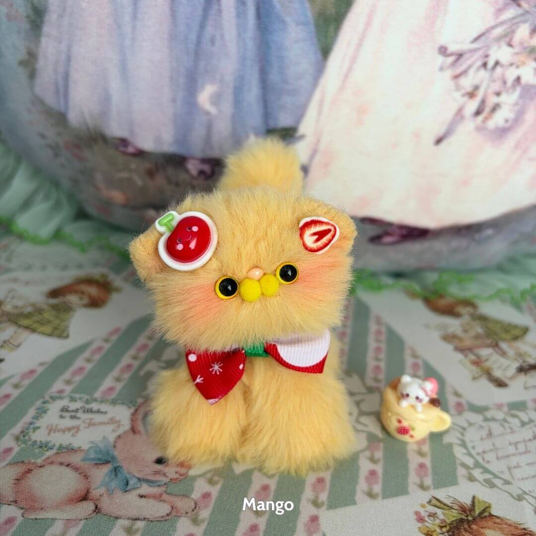 Cute miniature kitten in mango color with fruit-themed accessories, including a tomato hat and bow, from the Fruity Kitten Collection.
