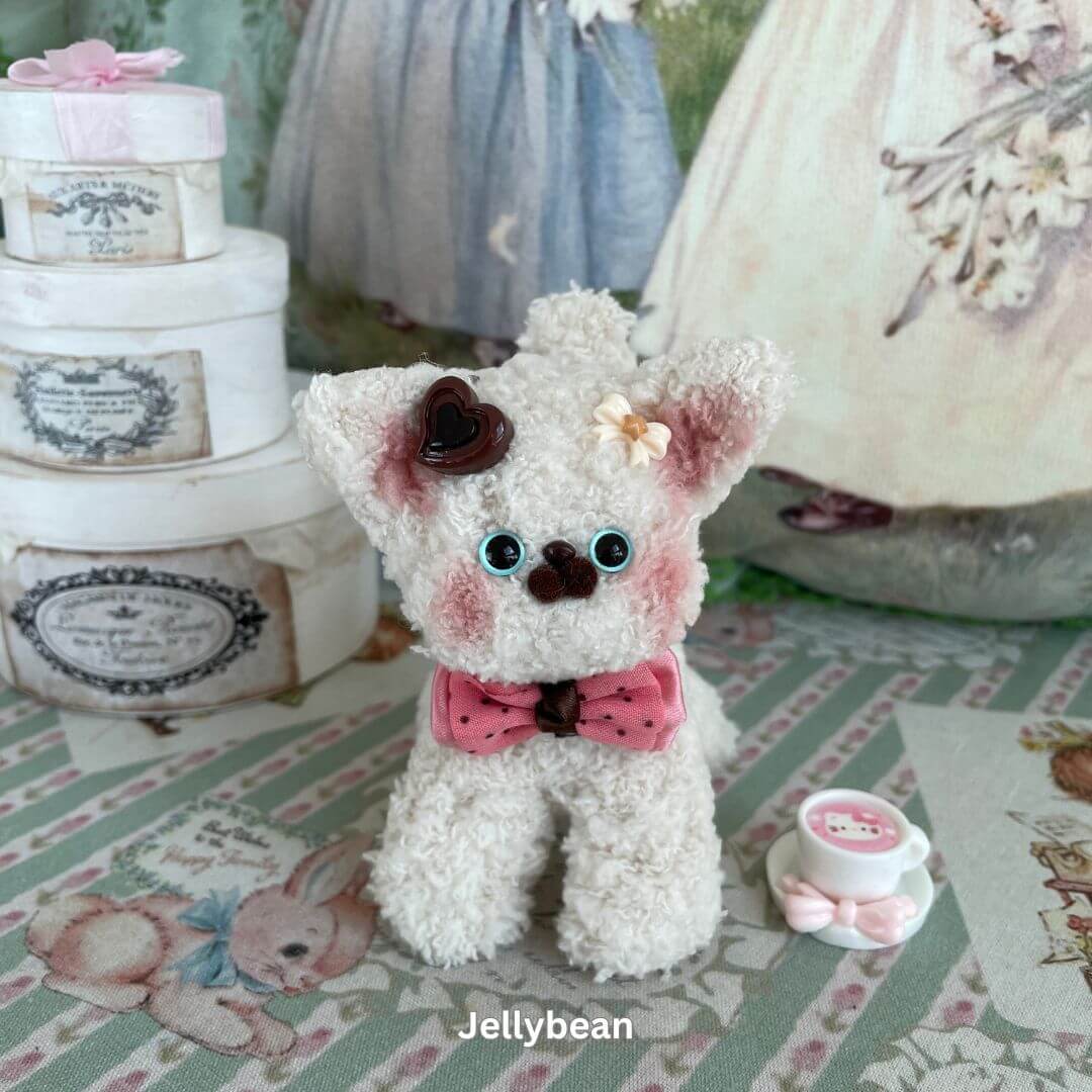 Handmade Devon Rex plush toy with a pink polka-dot bow, chocolate heart, and flower accessory, displayed on a decorative background.