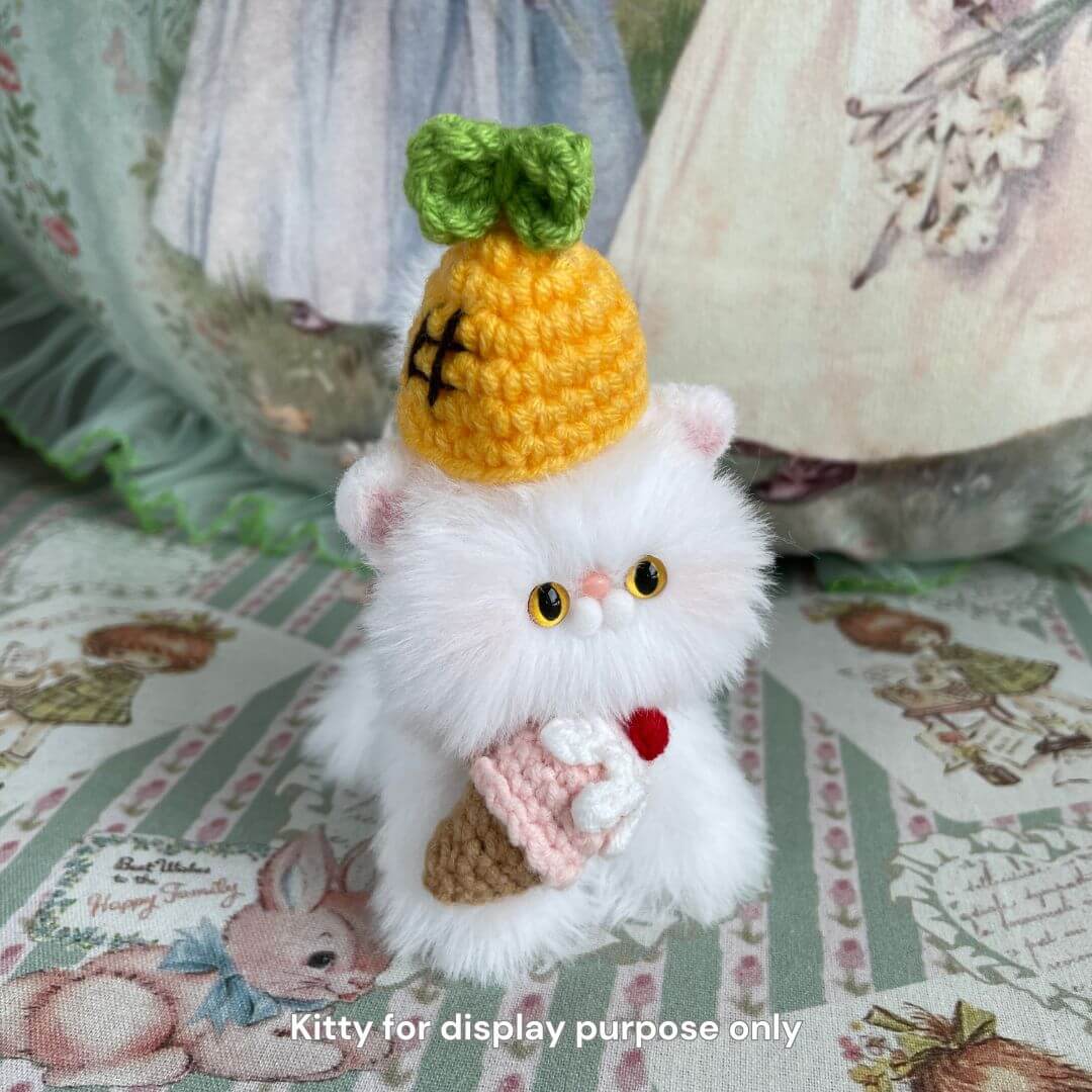 Cute plush kitten toy wearing a handmade pineapple hat and holding a tiny crochet cupcake on a decorative surface.