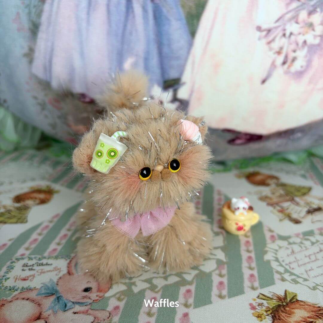 Handmade kitten from the Twinkling Kitten Collection, fluffy beige with silver accents, wearing a pink bow and holding a green drink.