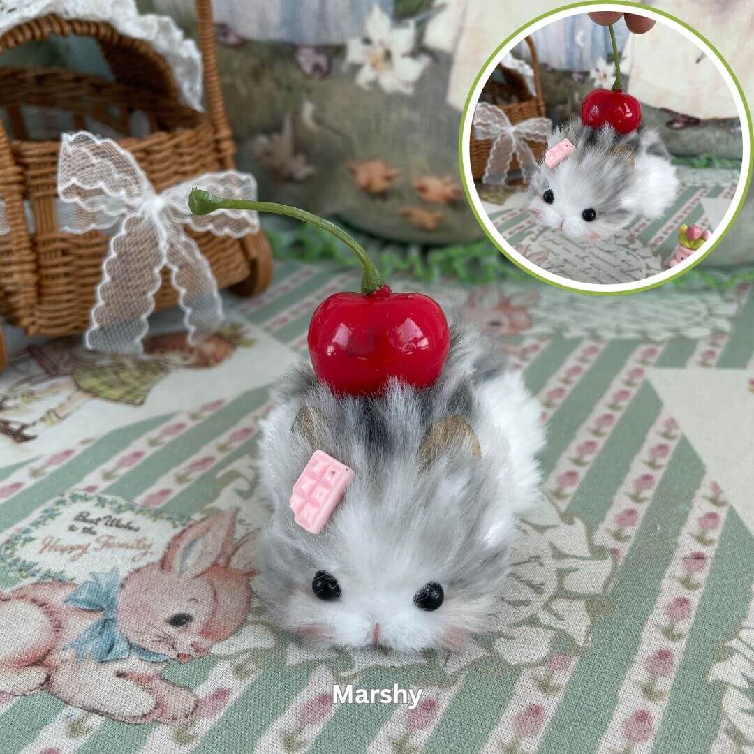 Plush hamster figurine with cherry and pink candy details, laying on a floral pattern, next to a woven basket and plush rabbit.