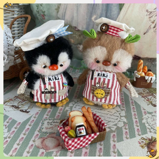 Two handmade mini chef penguins in aprons and hats, with a bread basket, on a decorated table cloth.
