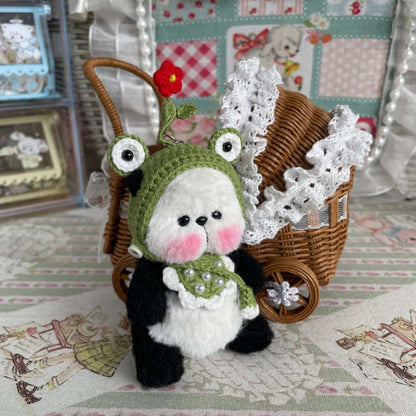 Hand-crocheted mini panda with movable joints wearing a green hat and apron, next to a decorative wicker stroller, handmade by René.