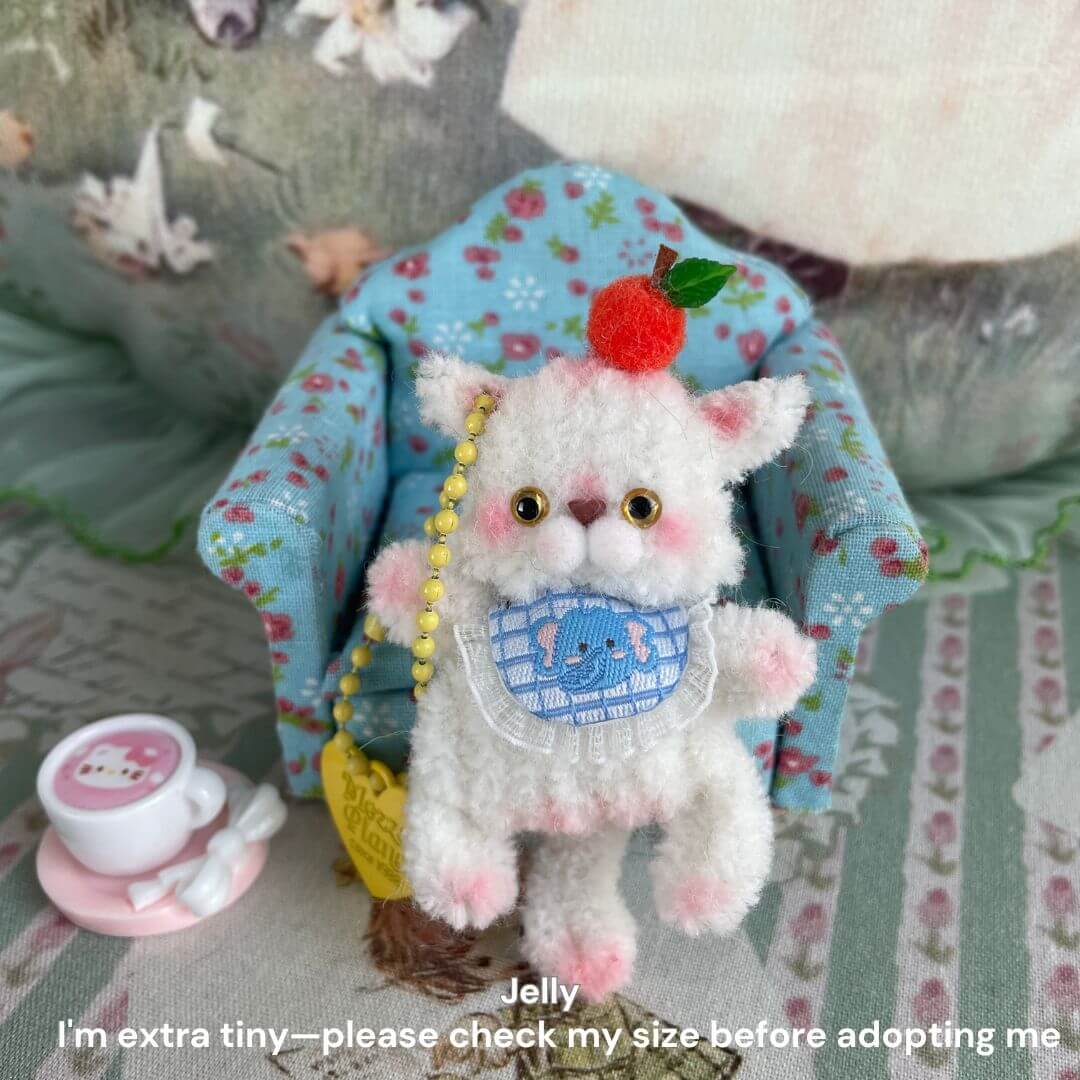 Miniature plush kitten with cherry hat and bib, sitting on floral chair, next to cute tea set; perfect accessory for bags or desks.
