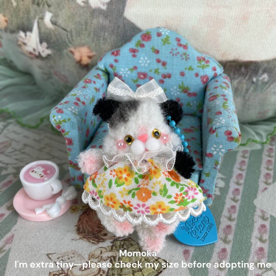 Miniature handmade cat plush toy in floral dress sitting on a blue patterned chair with tiny tea set, part of the Pipe Cleaner Animals Collection