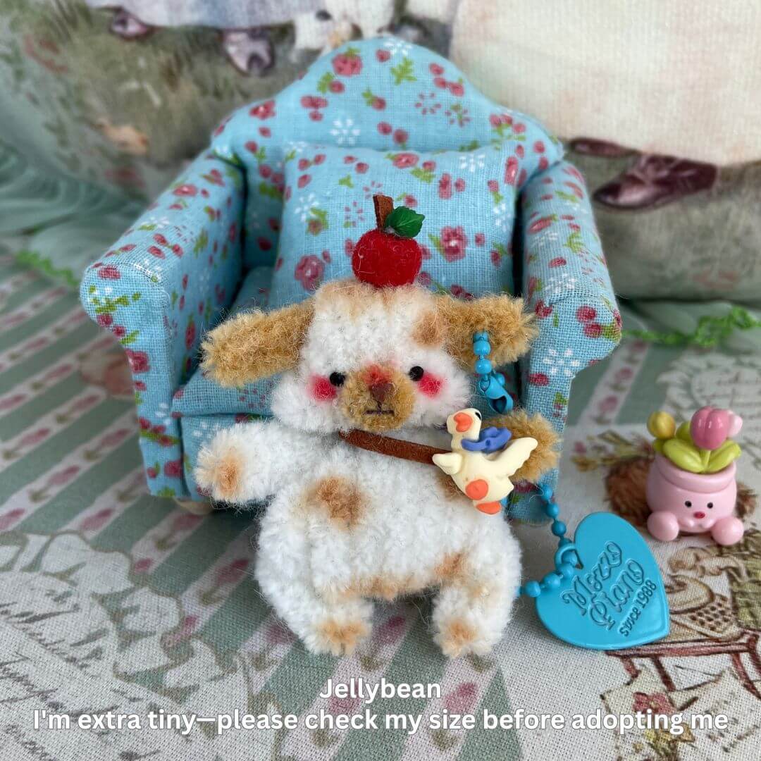 Miniature plush puppy with cherry hat and beaded keychain, sitting on a floral armchair.