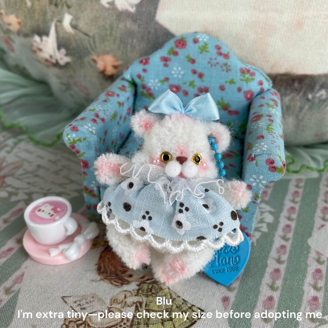 Tiny plush animal with blue dress and bow, sitting on a floral chair with pink cup, featuring delicate details, labeled 'Blu'.