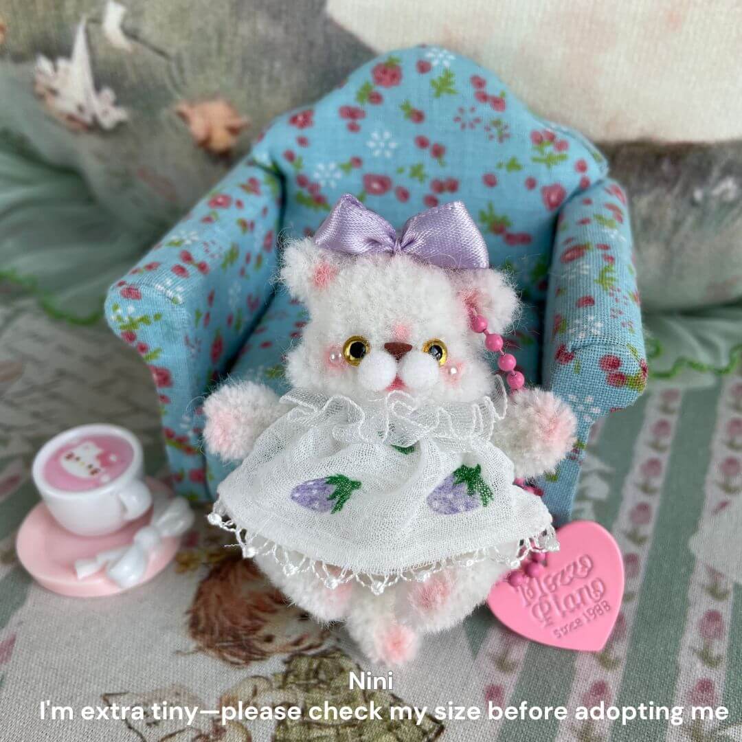 Miniature white plush kitten in floral chair with purple bow and apron, surrounded by tea set, featuring adoption reminder.