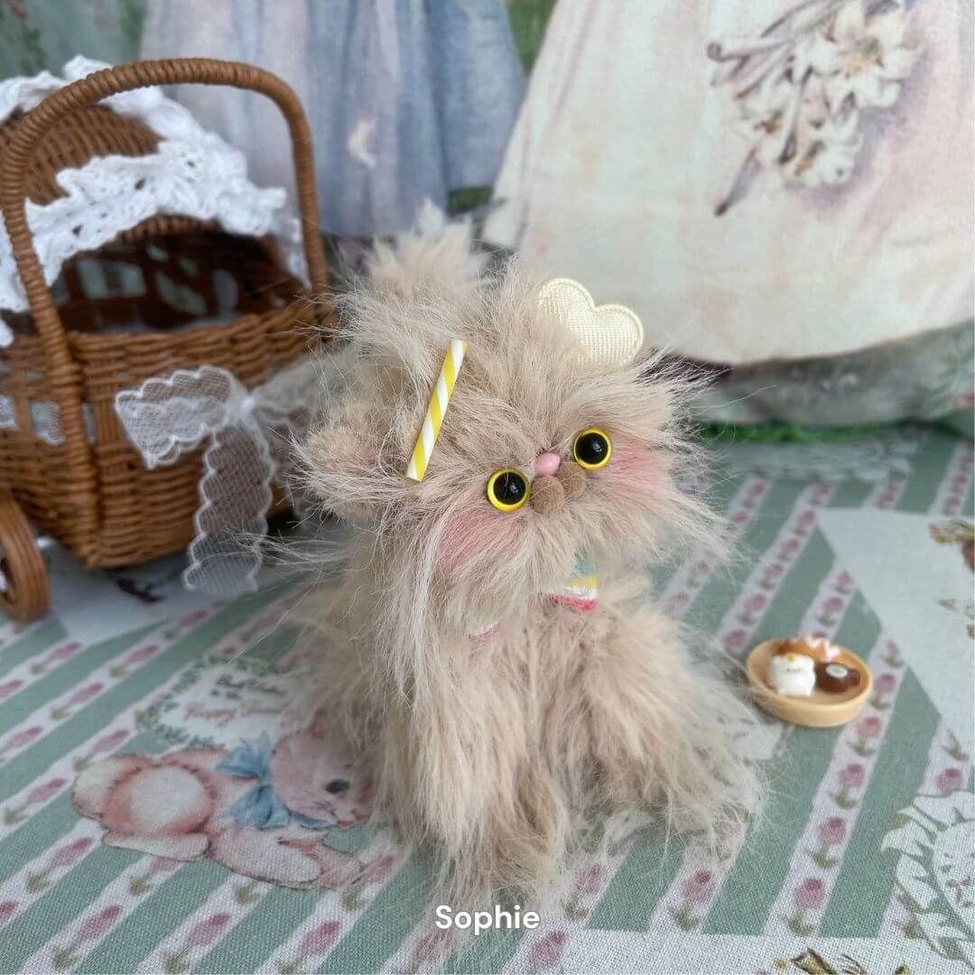 Handmade fluffy kitten toy from Siberian Snuggles Collection with whimsical accessories on patterned textile background.