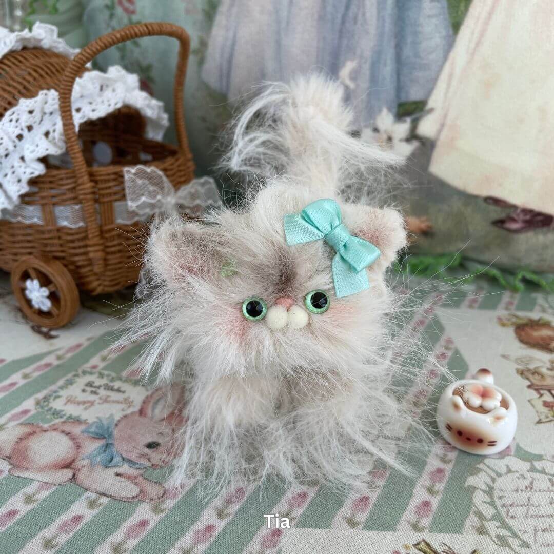 Handmade fluffy kitten plush toy with blue bow from the Siberian Snuggles Collection, set on a vintage patterned fabric background.