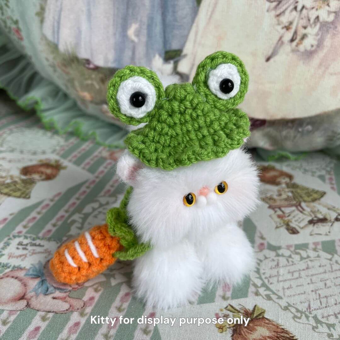 Handmade plush kitten wearing a green froggy hat and orange carrot sling, showcasing Snuggle Bits accessories for pets.