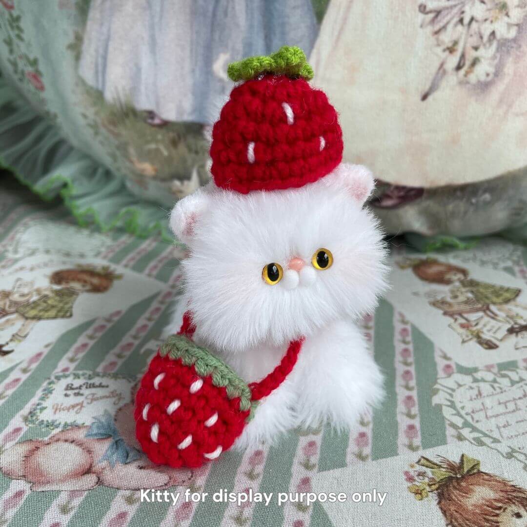 Fluffy white plush cat wearing a red strawberry hat and bag, sitting on a floral fabric backdrop. Perfect accessory for plush toys.