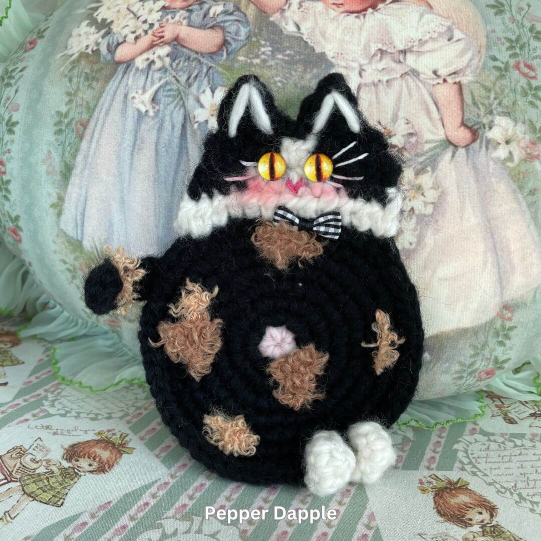 Handmade plush black cat toy with orange patches, featuring bright yellow eyes, set against a vintage-style floral background.